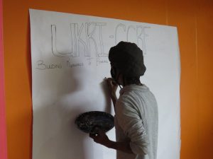 A person stands facing a large sheet of paer on an orange wall. Their back is to the camera. They hold a pen or paint brush and a palette.